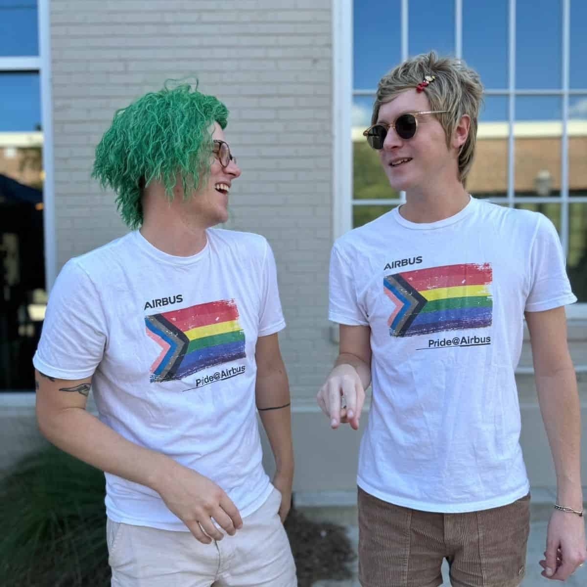 Two people with colorful hairstyles wearing matching shirts with a rainbow design laugh together in front of a brick wall.