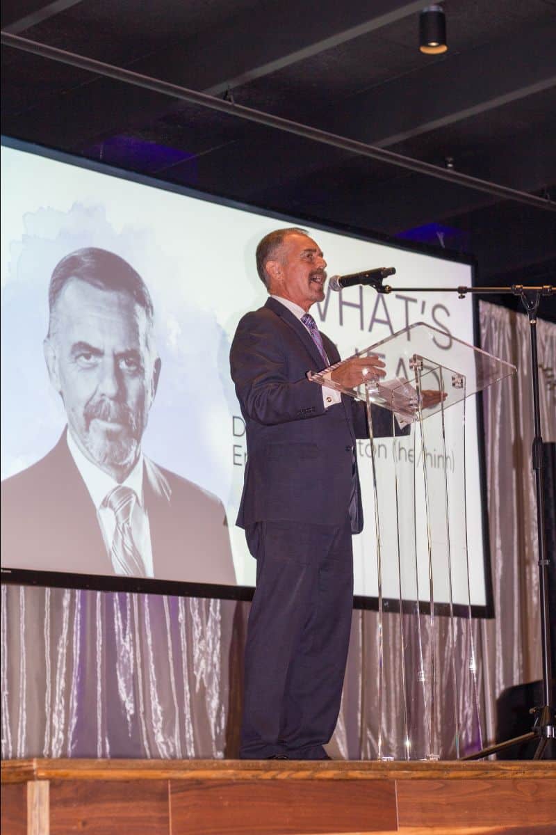 A man in a suit speaks at a podium in front of a screen displaying another man's portrait.