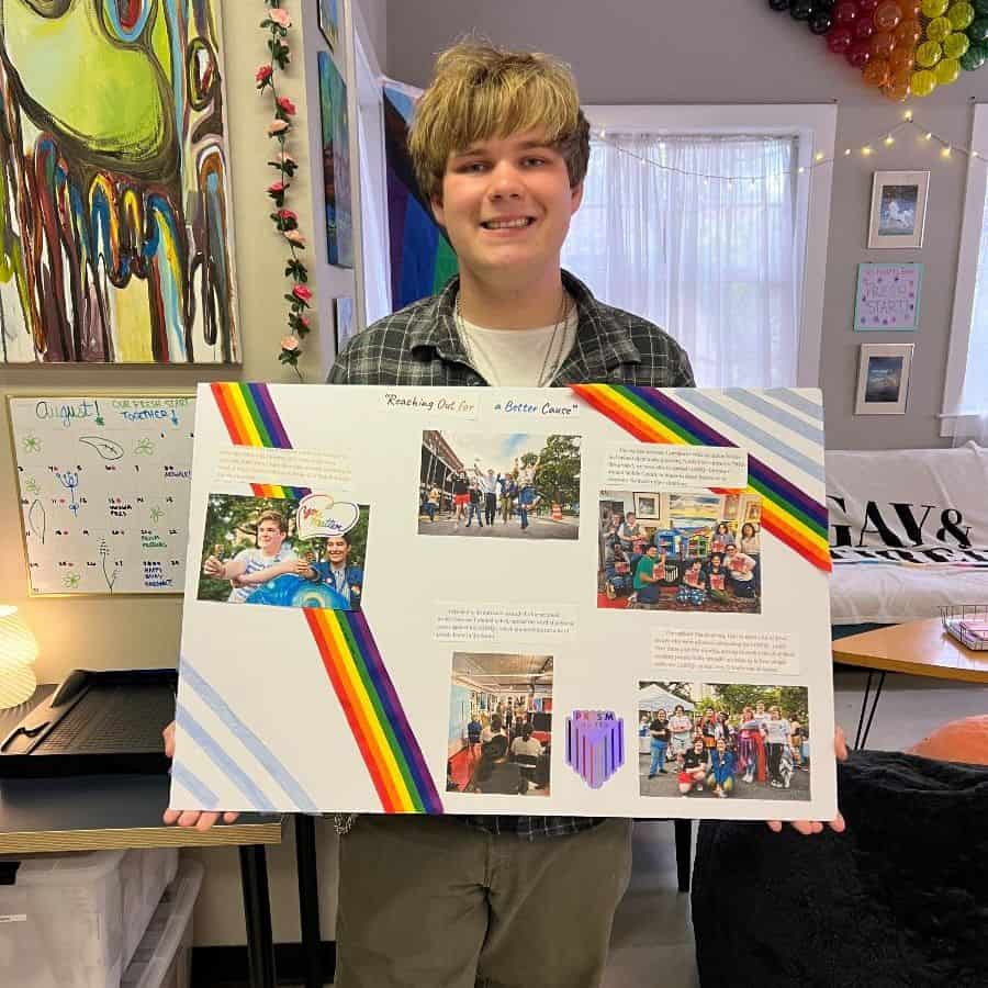 A person stands indoors holding a presentation board decorated with rainbow stripes and photos, with a colorful and cozy room in the background.