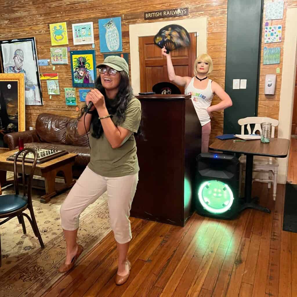 A person sings into a microphone in a casual indoor setting, while another person, wearing a blonde wig and holding a fan, stands beside a speaker in the background. The room has various artworks on the walls.