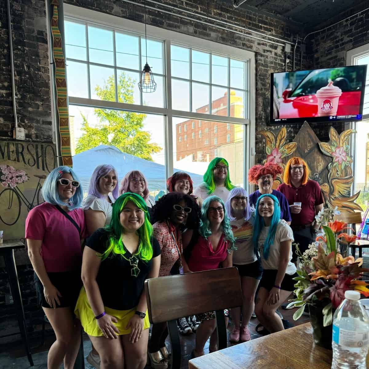 Group of people with colorful wigs posing indoors near large windows, with a TV screen and floral arrangement in the background.