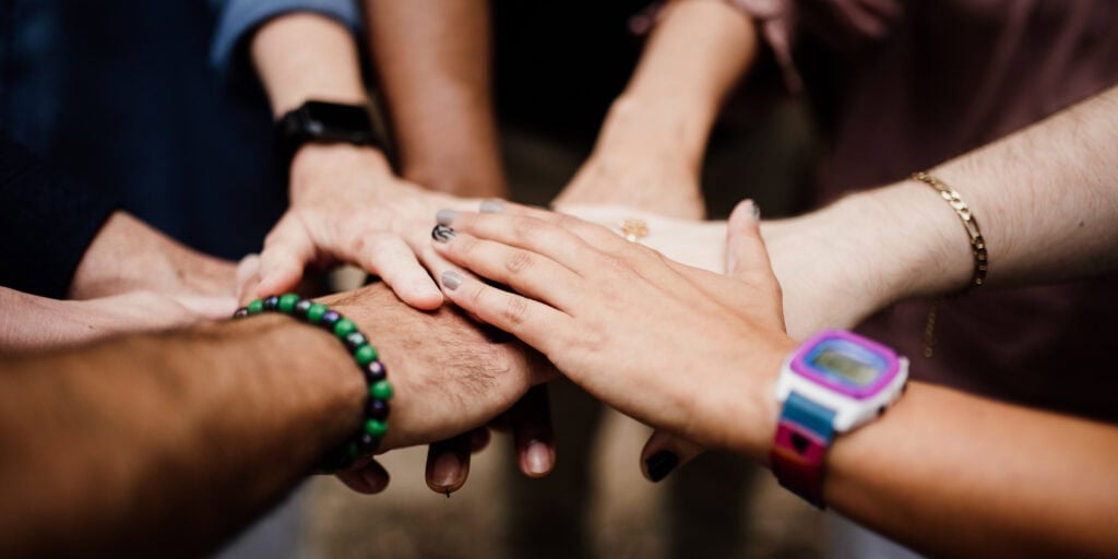 A group of six people place their hands together in a stack, showing teamwork and unity. Various skin tones, jewelry, and watches are visible.