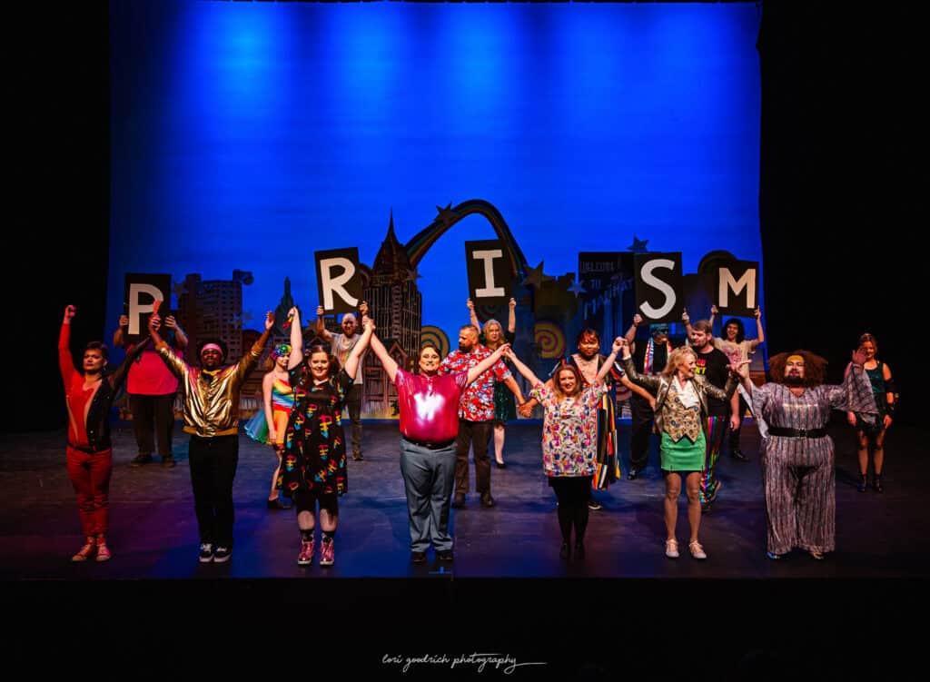 A diverse group of performers stand on stage holding hands with their arms raised in front of a backdrop with cityscape artwork and large letters spelling "PRISM.