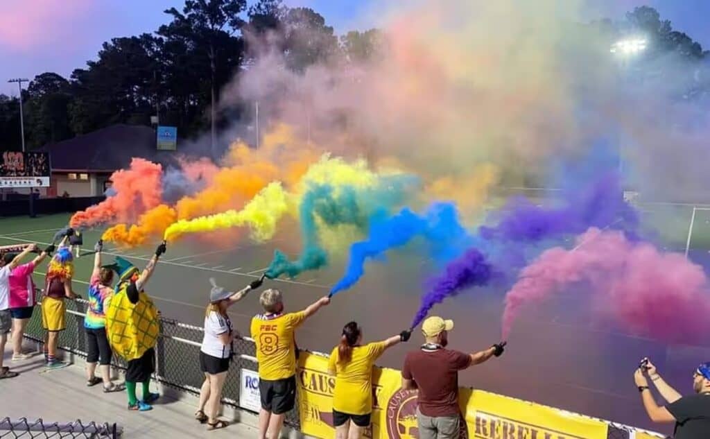 A group of people at a sports stadium holds colorful smoke flares, creating a rainbow smoke effect over the field.