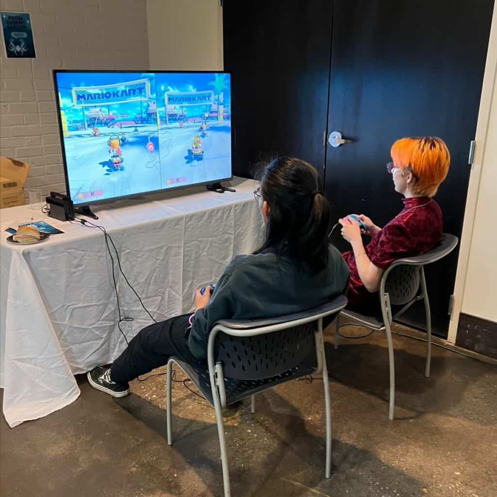 Two people playing Mario Kart on a TV in a casual indoor setting, seated on chairs, engaged in the game.