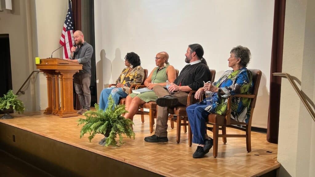 A person speaks at a podium while five others sit on chairs on a stage. An American flag is in the background.