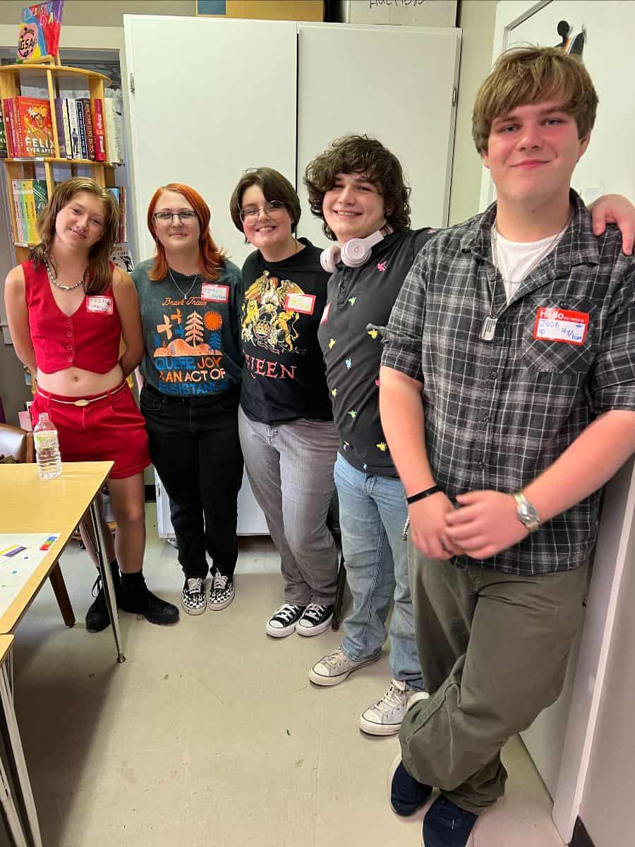 Five teenagers with name tags stand and smile in a classroom near bookshelves and cabinets. One wears headphones, another holds a water bottle on a desk.