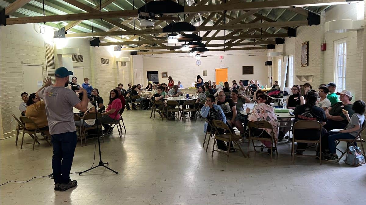 A man speaks into a microphone at the front of a large, well-lit room filled with people seated around round tables, listening attentively.