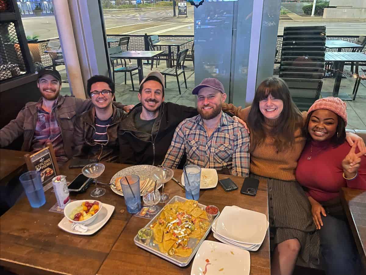 A group of six people sitting at a restaurant table with plates of food and drinks in front of them. They are smiling at the camera.