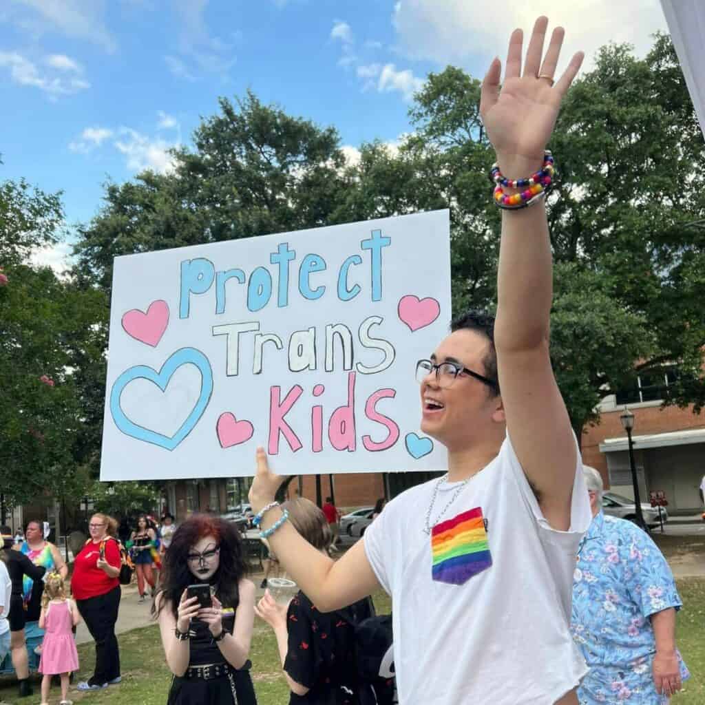 A person raises their hand while holding a sign that reads "Protect Trans Kids" at an outdoor gathering. Others are seen in the background. 