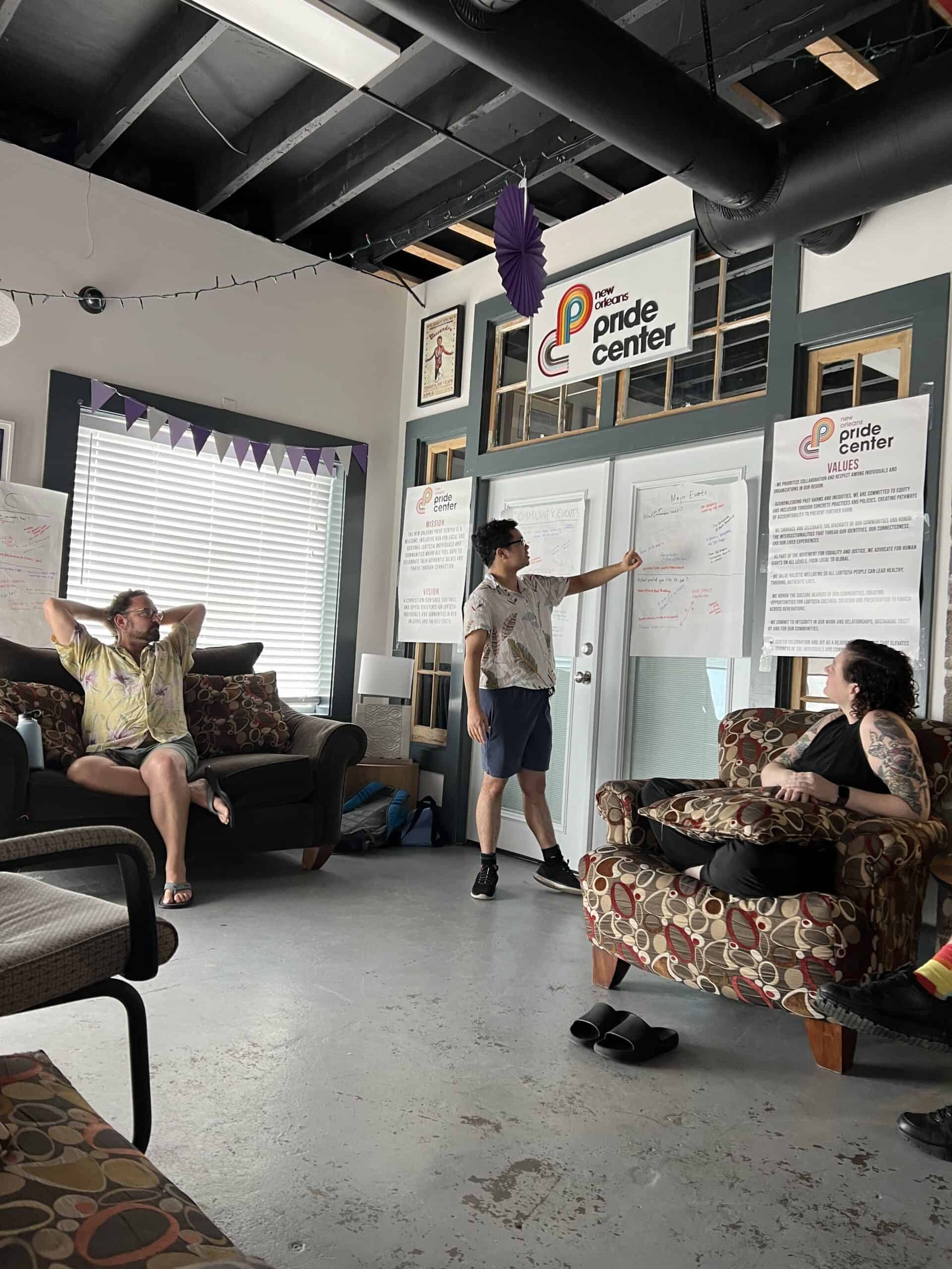 Three people are in a brightly lit room at the Pride Center; one person stands and points to notes on large sheets of paper, while the others sit and listen.