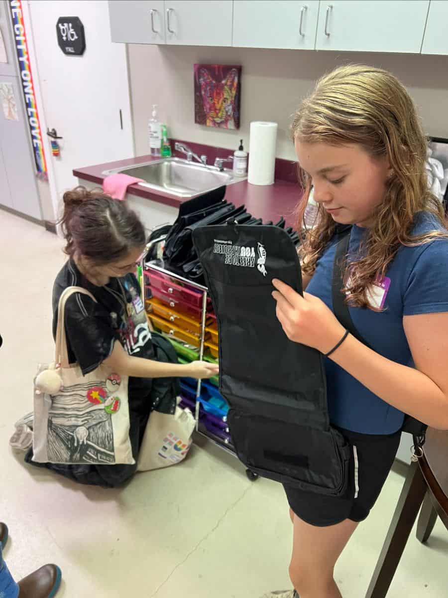Two girls are in an art classroom; one is kneeling and reaching into a colorful storage cart, while the other holds and looks at a black folded apron or bag.
