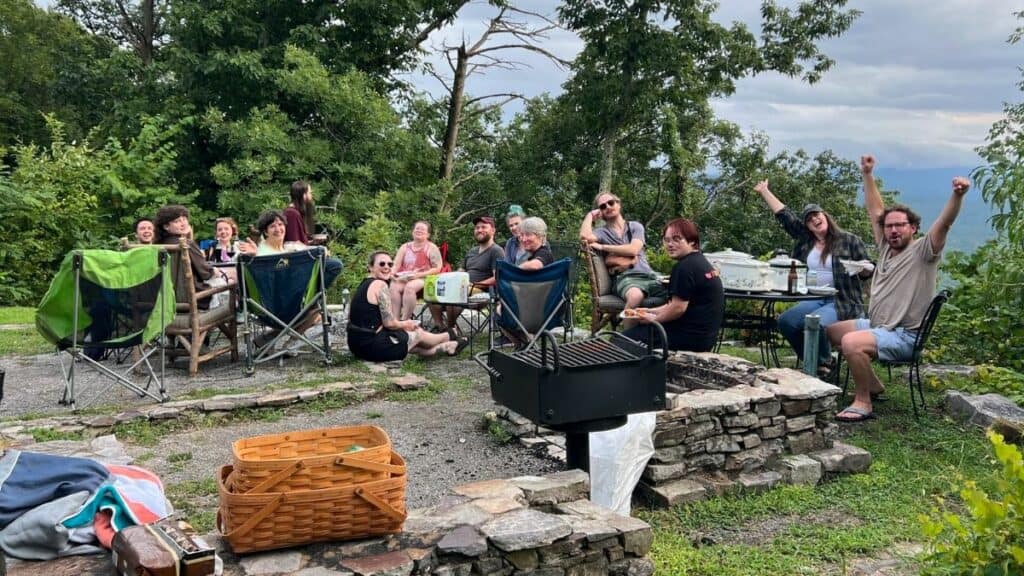 A group of people sitting outdoors around a table and in chairs, some are raising their arms, with a grill and picnic basket in the foreground. Trees and a scenic background are visible.