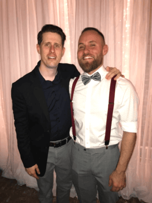 Two men stand side by side in front of sheer curtains with string lights, one wearing a dark blazer and the other in suspenders and a bow tie.