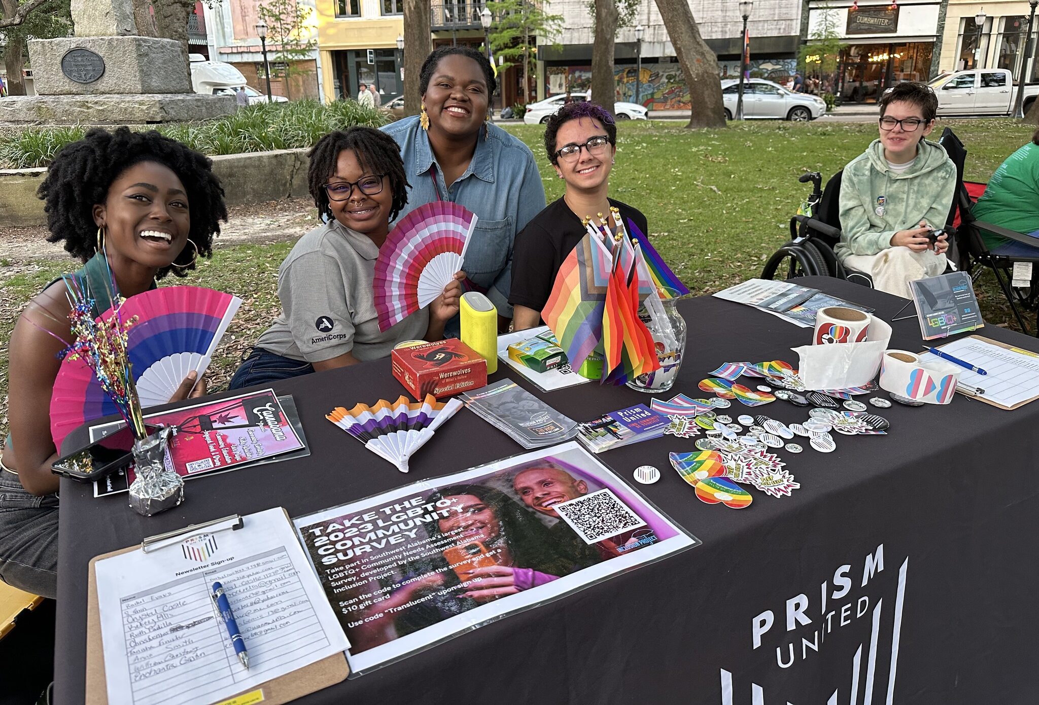 Five people sit at an outdoor table covered with LGBTQ+ flags, pamphlets, and buttons for Prism United, with survey forms and informational materials displayed.
