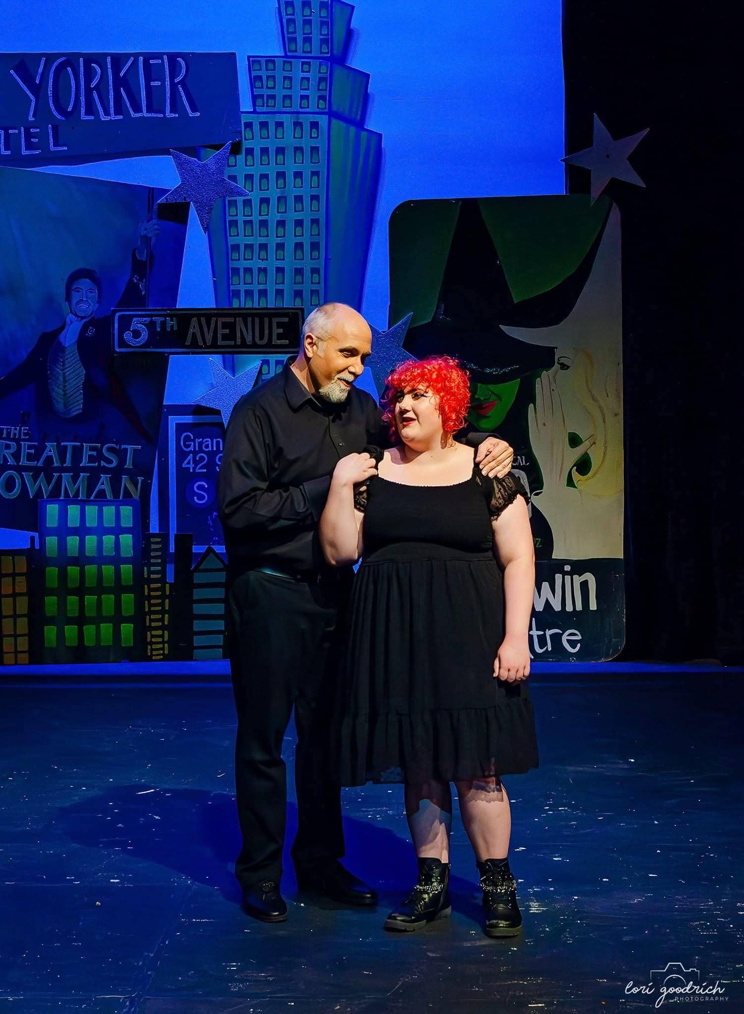 Two people in black outfits stand on a theater stage with cityscape and musical posters as the backdrop, under bright stage lighting.