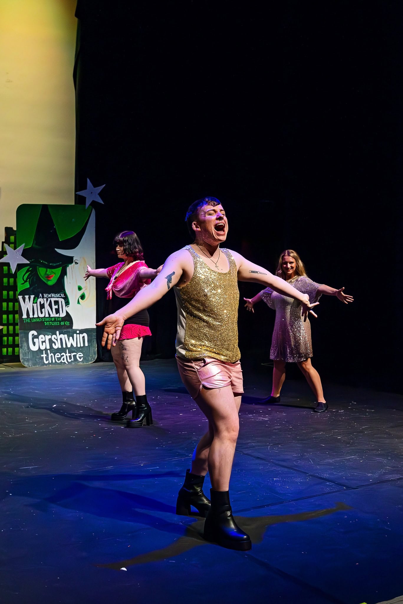 Three people perform on stage in colorful outfits; one stands in front with arms wide, and a "Wicked Gershwin Theatre" sign is visible in the background.