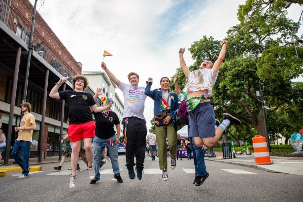 Five people holding small rainbow flags jump in the air on a city street during an outdoor event, with buildings, trees, and other participants visible in the background.