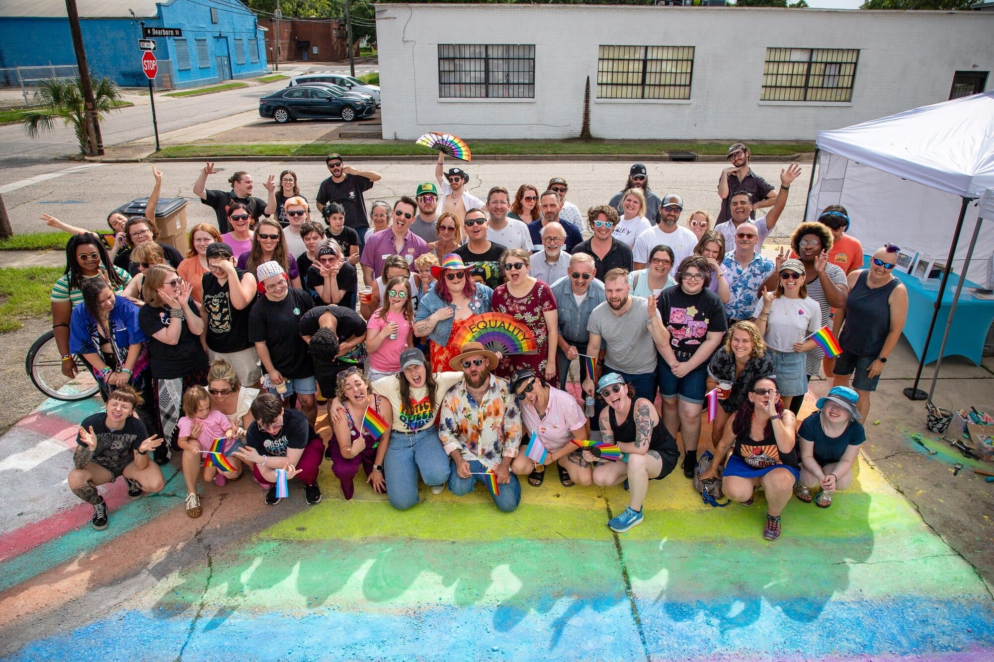 A large group of people poses outdoors on a colorful painted crosswalk, some holding rainbow flags and wearing Pride-themed clothing, smiling and waving at the camera.
