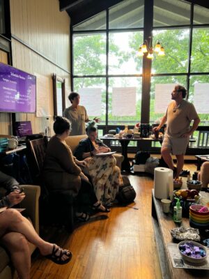 A group of people having a casual meeting in a room with large windows. The person standing near a screen appears to be speaking while others are seated and listening. Notes are posted on the windows.