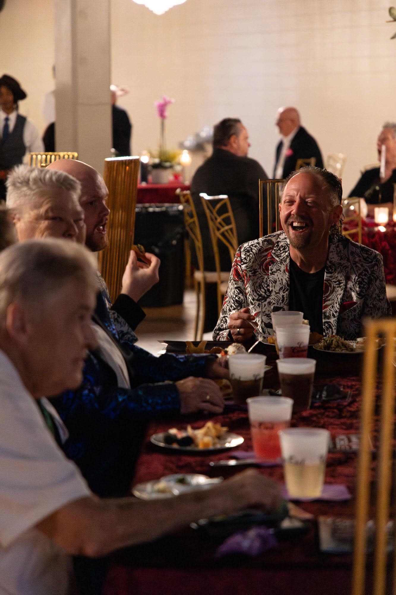 Four people sit at a table with food and drinks, engaging in conversation and laughter at a formal indoor event. Other guests are visible in the background.