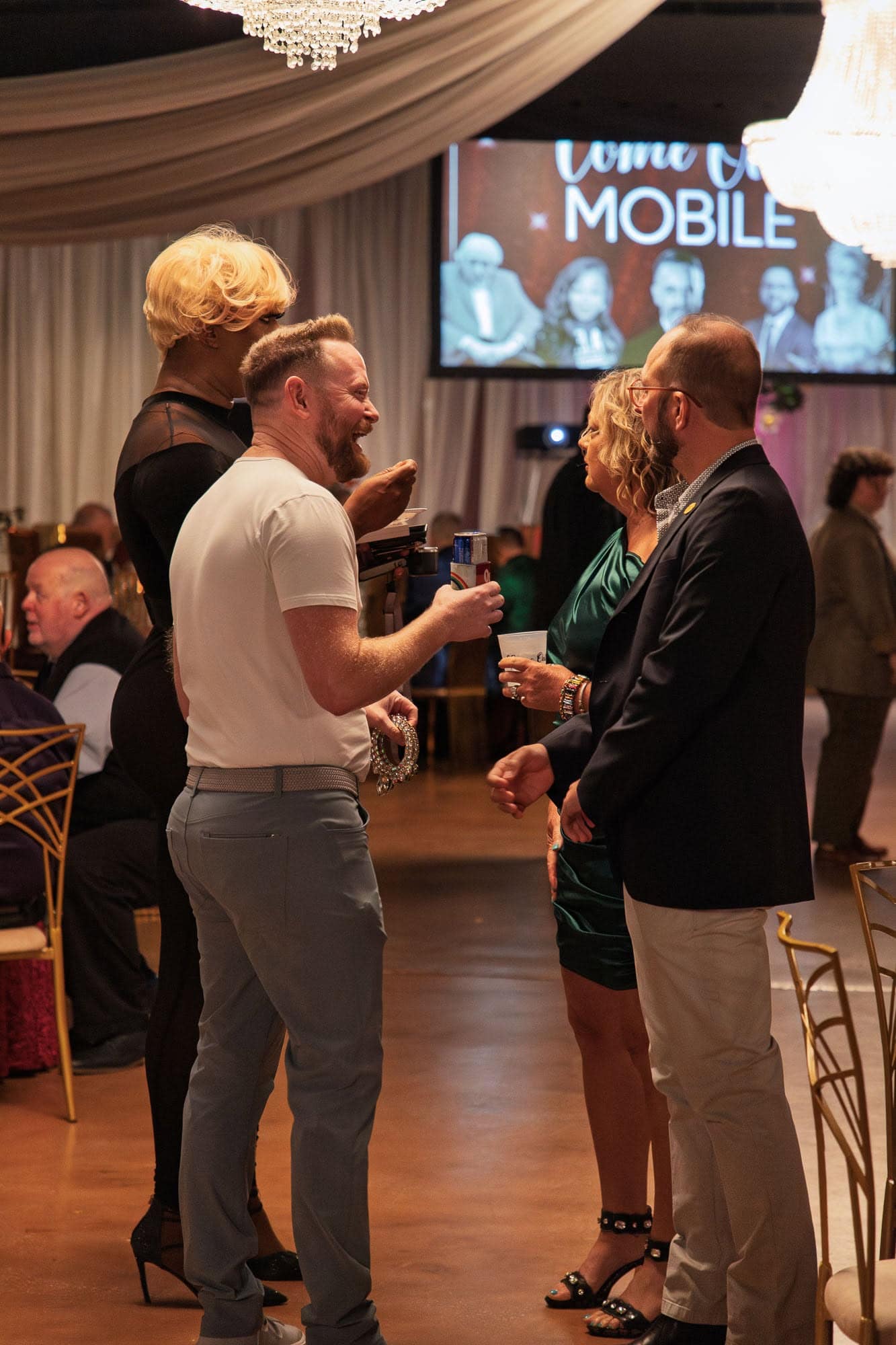 Four adults stand in a group, conversing and holding drinks at an indoor event with formal decor; a screen with the word "MOBILE" is visible in the background.