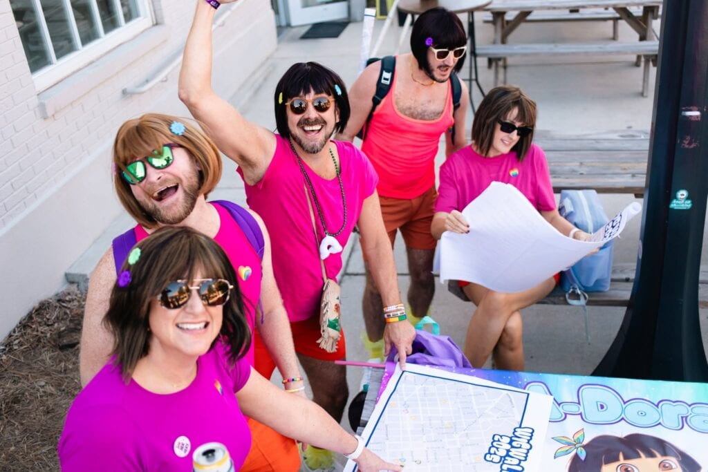 Five adults in brightly colored outfits and wigs stand outside near a table with a map and board game, appearing cheerful and playful.