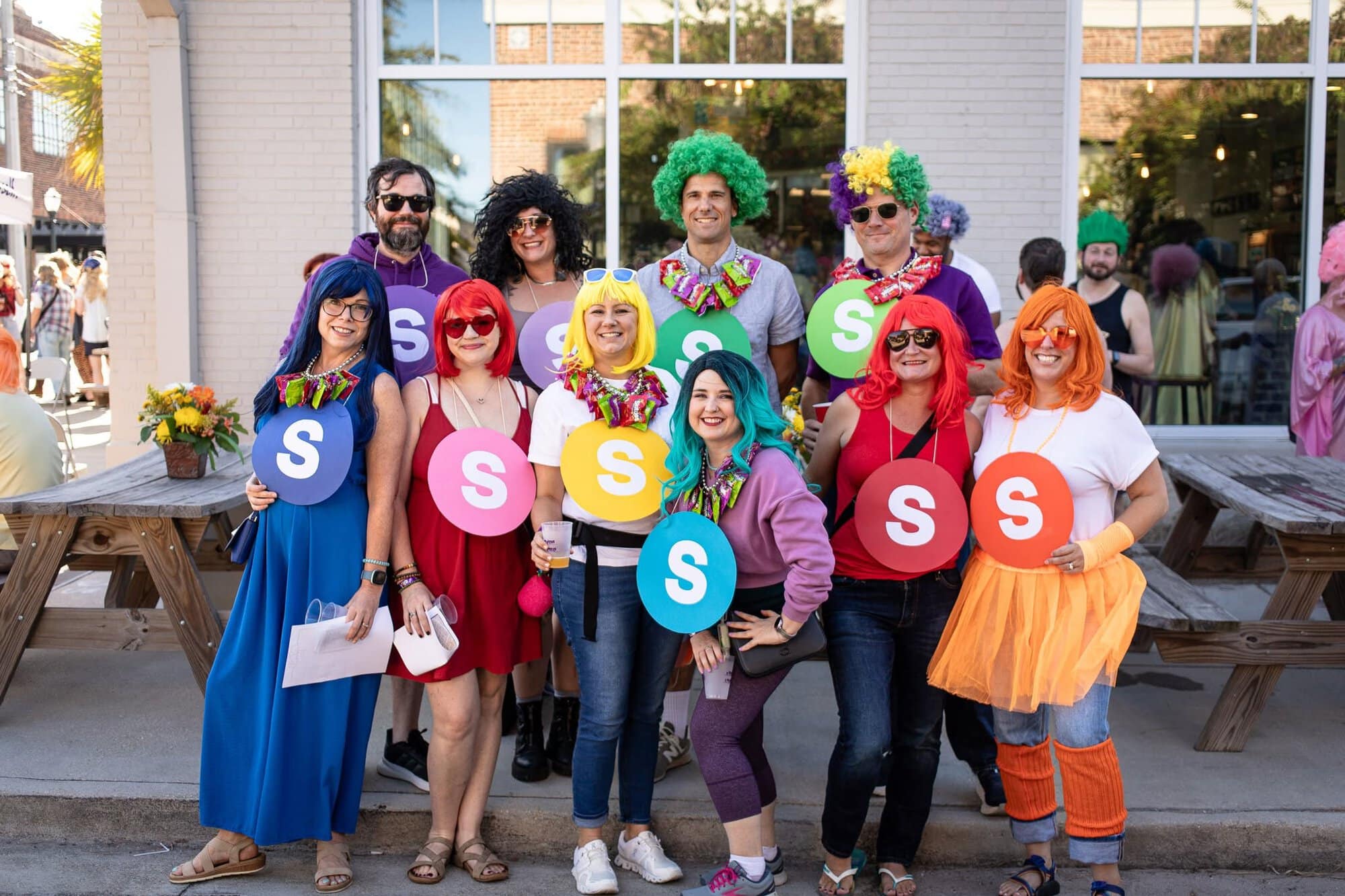 A group of people in colorful wigs and costumes pose outdoors, each holding a round sign with the letter “S” on it.