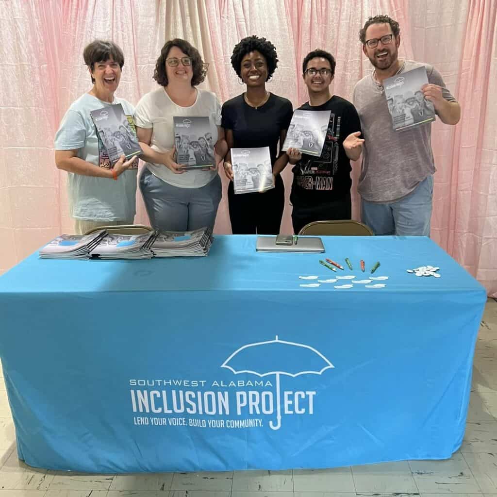 Five people stand smiling behind a table covered with a Southwest Alabama Inclusion Project cloth, holding up booklets.