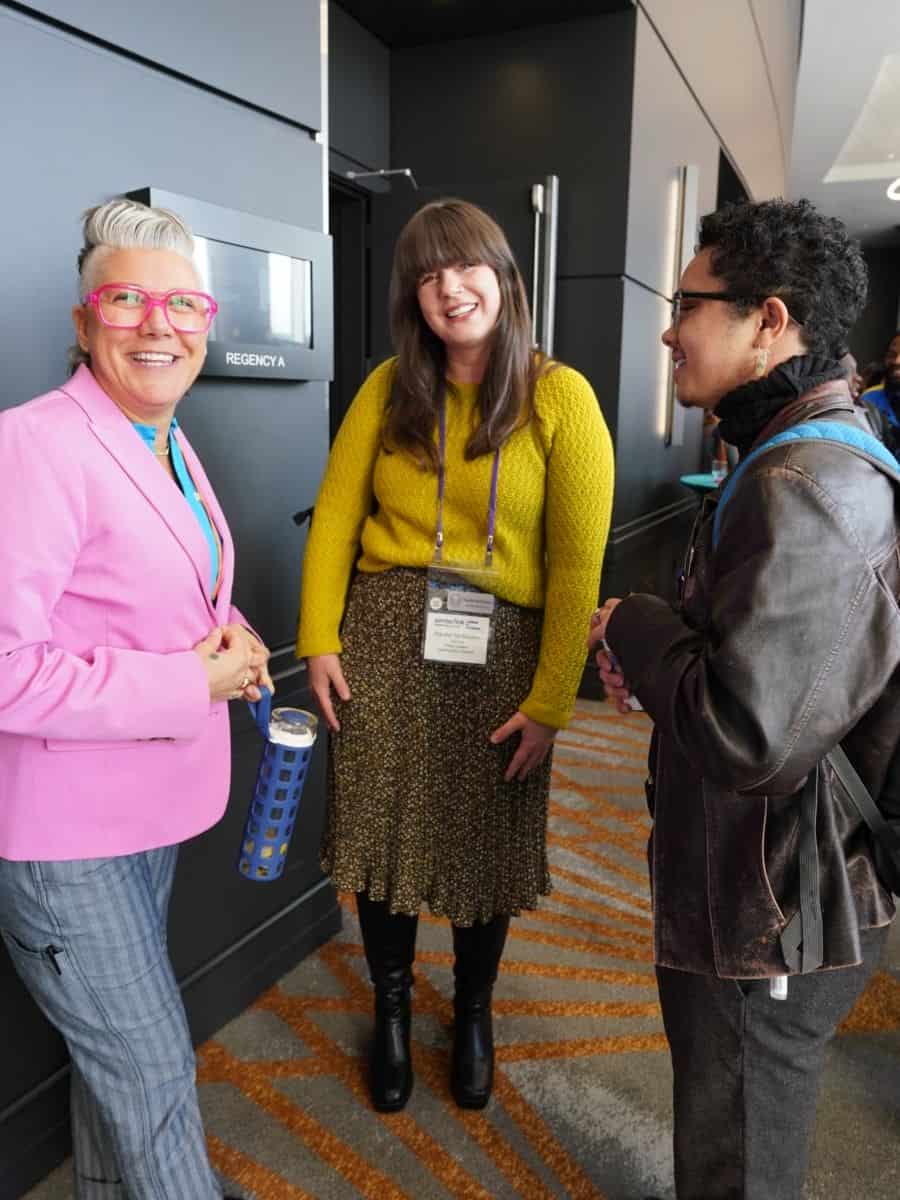 Three people in conversation in a hallway. One wears a pink blazer, another a yellow sweater, and the third a leather jacket. They appear to be at an event or conference.