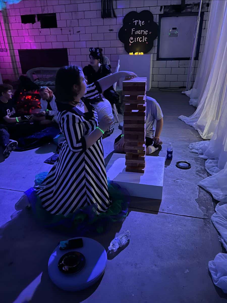 Person playing Jenga in a dimly lit room with colorful lights. A sign reads "The Faerie Circle" in the background.