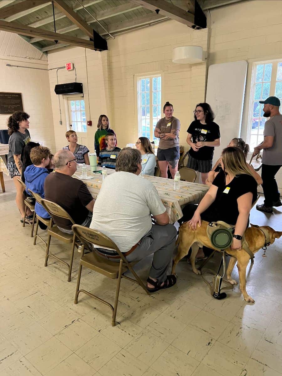 A group of people sit and stand around tables in a bright room, engaged in conversation; a dog sits beside one person.