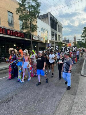 A group of people, some in colorful outfits, march down a city street holding a "PRISM" sign for a parade or event.