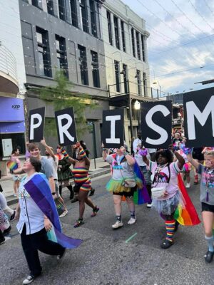 People walking in a street parade, carrying large signs that spell "PRISM." They are dressed in colorful, festive clothing and hold rainbow flags. Buildings and lights are visible in the background.