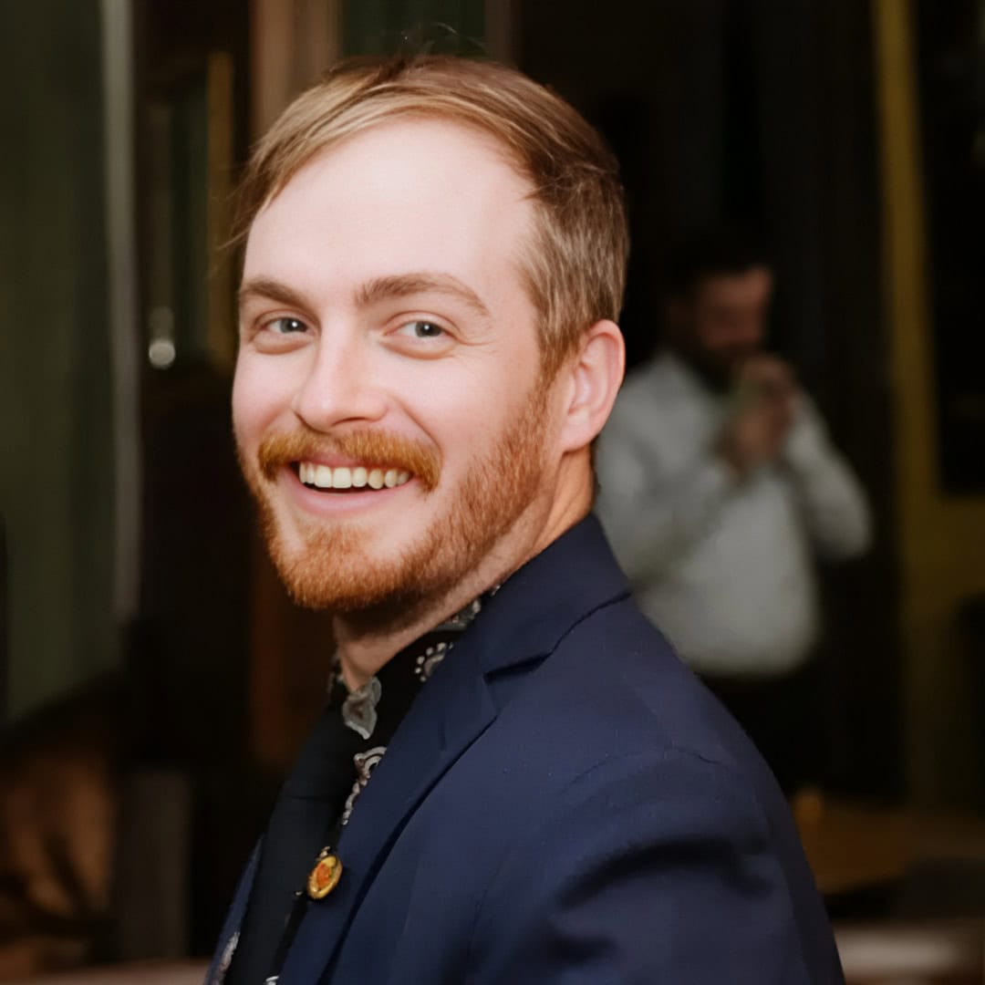 Smiling person with a beard, wearing a dark jacket, is looking over their shoulder indoors.