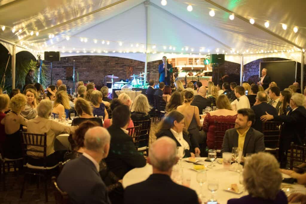 An evening event with guests seated at tables under a tent, while a band performs on stage in the background.