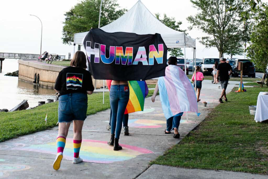 People are walking on a sidewalk near a body of water while holding and wearing various rainbow-themed items. One person holds a "HUMAN" sign adorned with rainbow colors.