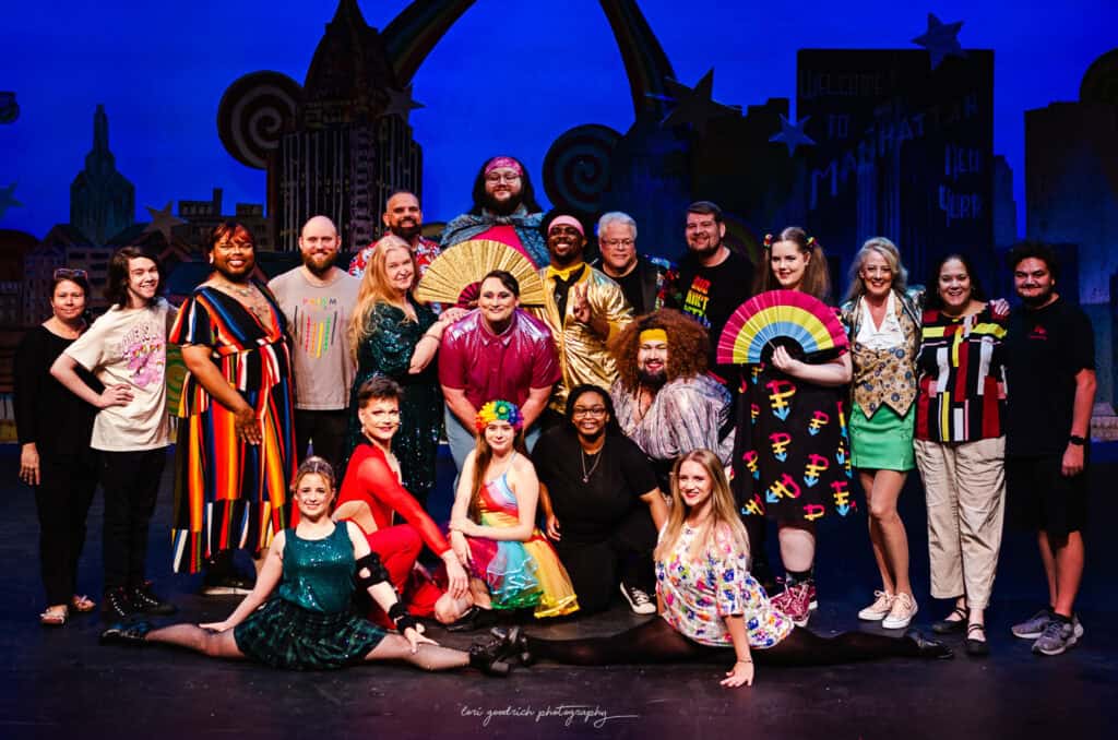 A group of people in colorful costumes posing on a stage with a city backdrop. Some hold large decorative fans. The atmosphere appears joyful and festive.