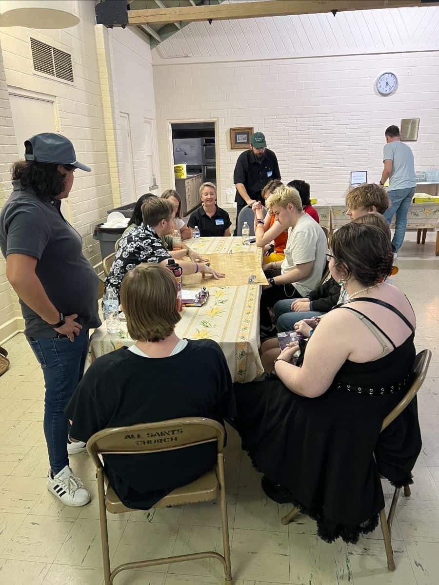 A group of people sit around a rectangular table inside a community room, engaging in a tabletop game; two people stand nearby.