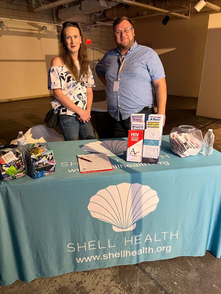 Two individuals stand behind a Shell Health table with brochures, a clipboard, and various items.