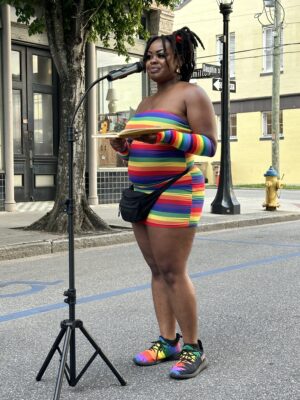 Person wearing a rainbow-striped outfit stands at a microphone in front of a building, holding a small tray.