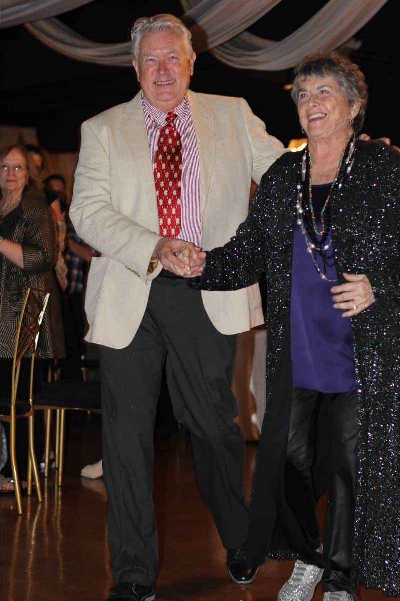 An elderly couple smiling and holding hands while walking, with the man wearing a beige jacket and the woman in a black sparkly outfit. Background shows seated guests and draped ceiling fabric.