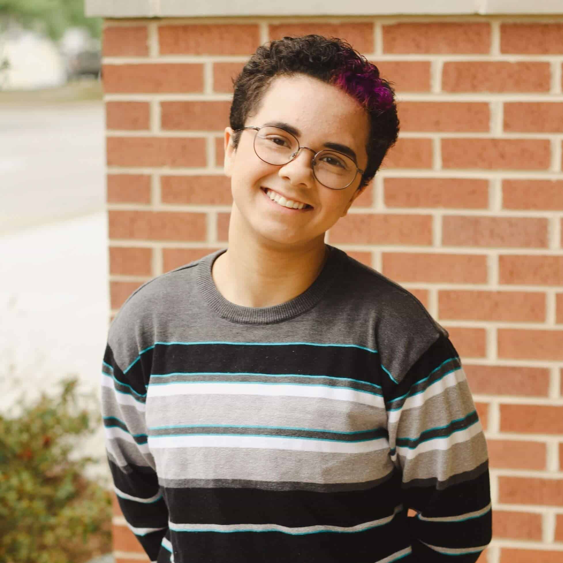 Person with glasses and a striped sweater smiling in front of a brick wall.
