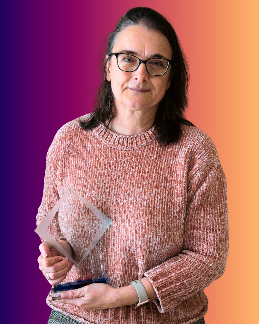 AngelaTrigg A person wearing glasses and a pink sweater holds a geometric glass award in front of an orange and purple gradient background.