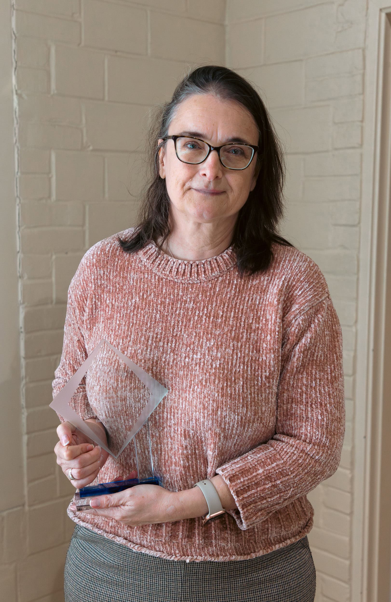 AngelaTrigg A woman wearing glasses and a pink sweater holds a translucent geometric award and a blue pen, standing against a light-colored brick wall.