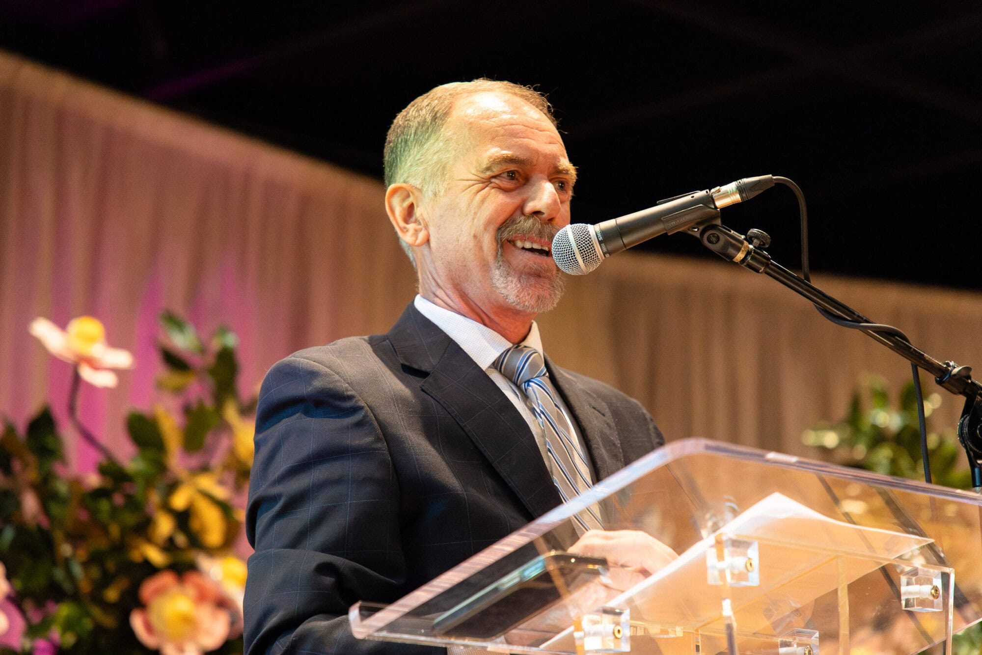 A man in a suit speaks into a microphone at a clear podium, with flowers and draped curtains in the background.