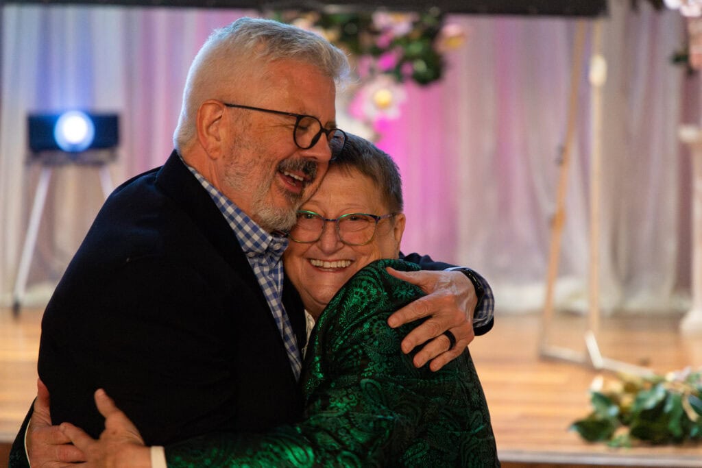 Two older adults wearing glasses hug and smile at an indoor event with soft lighting and floral decorations in the background.