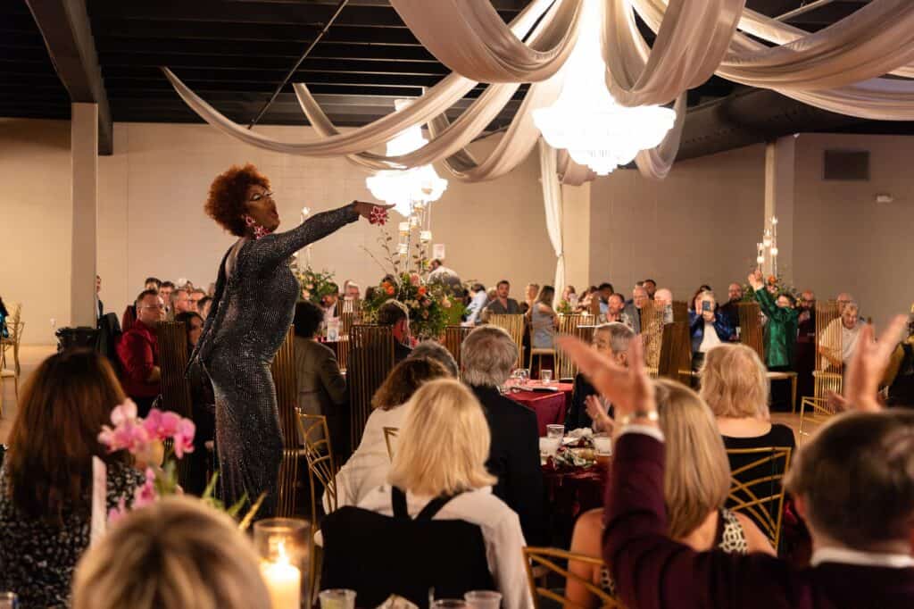 A performer in a sparkly dress interacts with a seated audience at a formal event in a decorated hall with draped fabric and chandeliers.