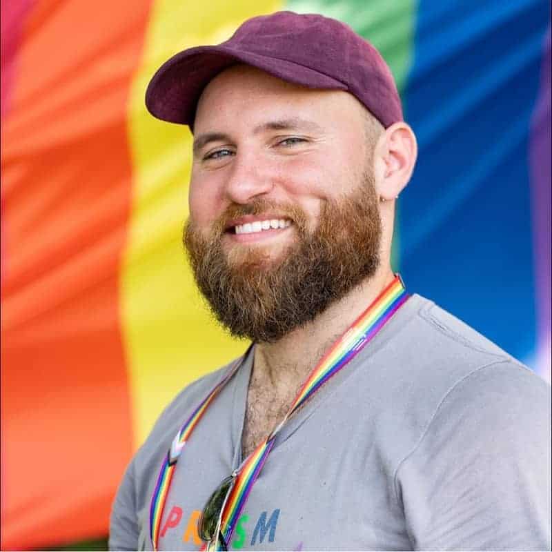 Man with a beard smiling in front of a rainbow flag.