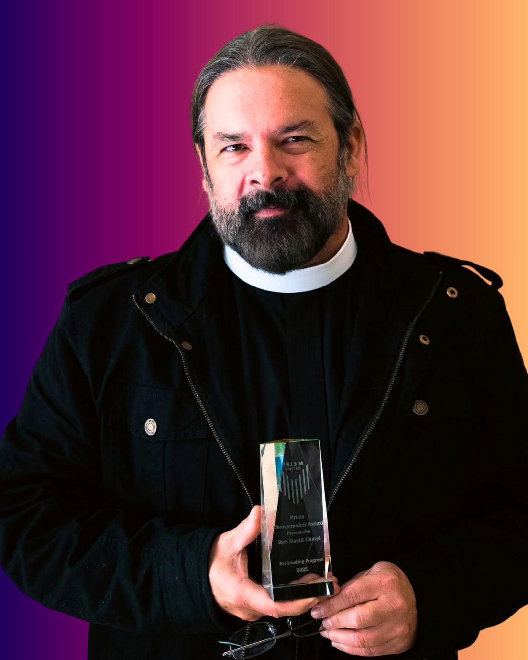 DaveChatel A person in a black jacket and clerical collar holds a clear award trophy and a pair of glasses, standing in front of a gradient background.